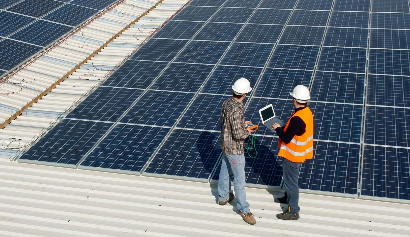 Two technicians inspecting solar panels on a rooftop, one holding a laptop and the other using a multimeter, showcasing solar panel services.