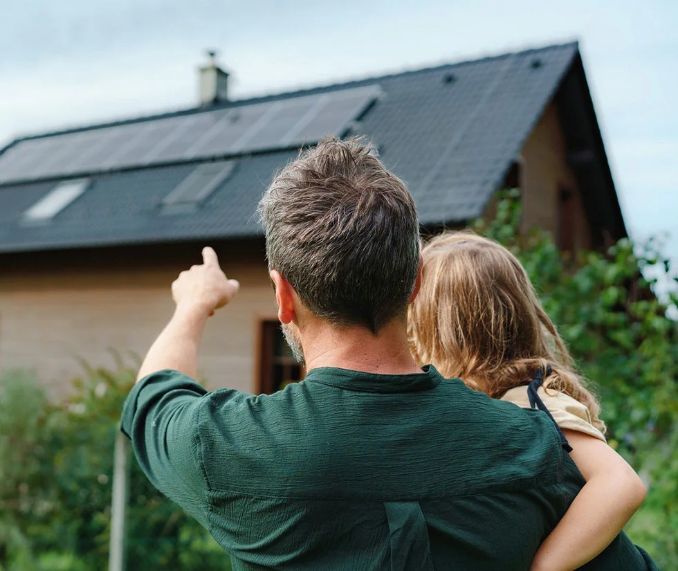 Man pointing at solar panels on a house roof while holding a child, illustrating family engagement with sustainable energy solutions.