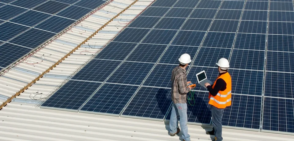 Two technicians inspecting solar panels on a rooftop, discussing data on a laptop, emphasizing innovative solar solutions and sustainable building practices.