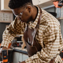 Man in plaid shirt and leather apron using a power drill in a workshop setting, demonstrating hands-on skills relevant to solar energy installations.