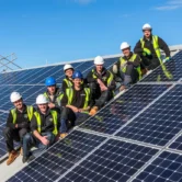 Group of solar panel installers wearing safety gear and hard hats, posing on a roof covered with solar panels, showcasing teamwork in sustainable energy solutions.