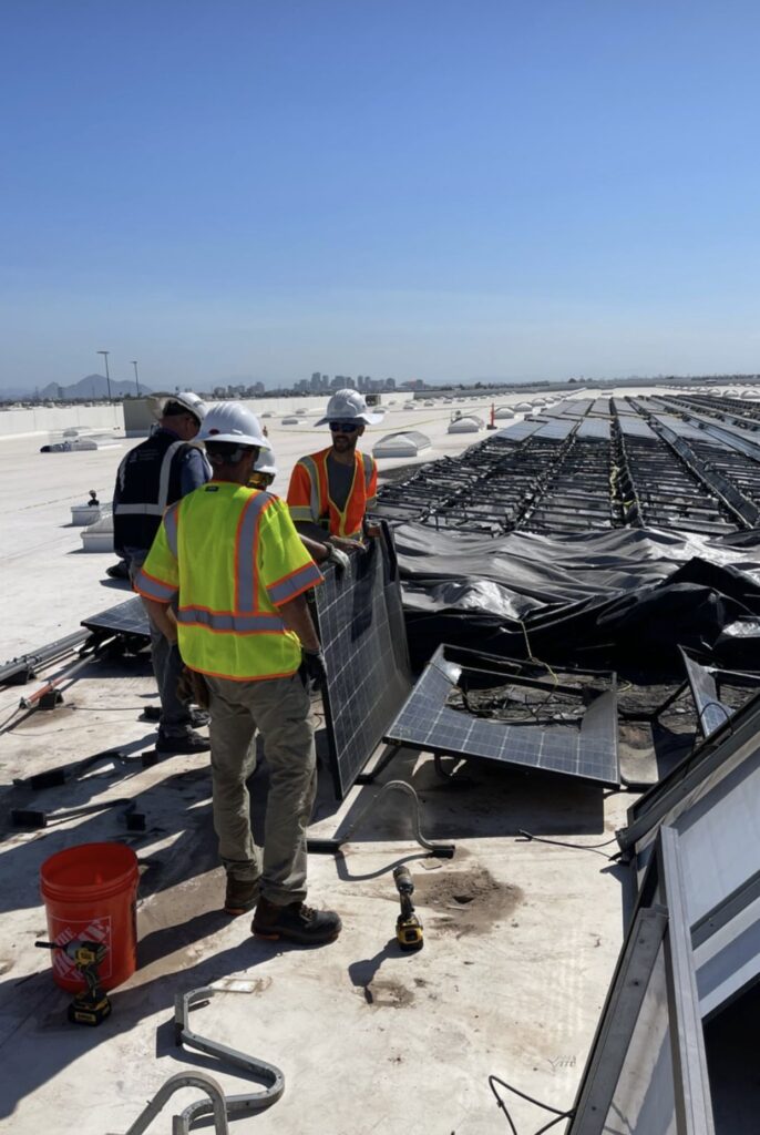 Technicians repairing solar panels on a rooftop, wearing safety gear and handling equipment, with city skyline in the background, reflecting professional solar system repair services in Arizona.